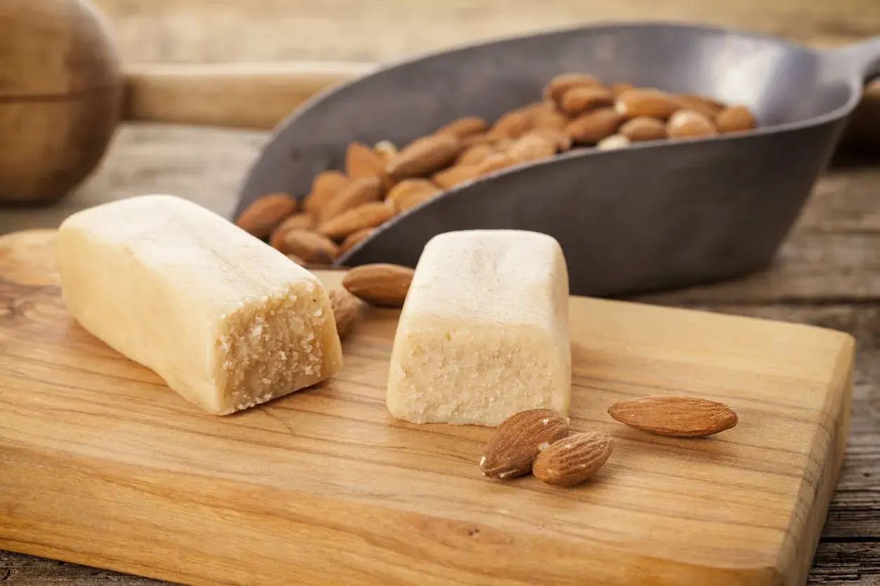 Deux blocs de pâte d'amande maison sur une planche en bois, avec des amandes dans une pelle en arrière-plan.