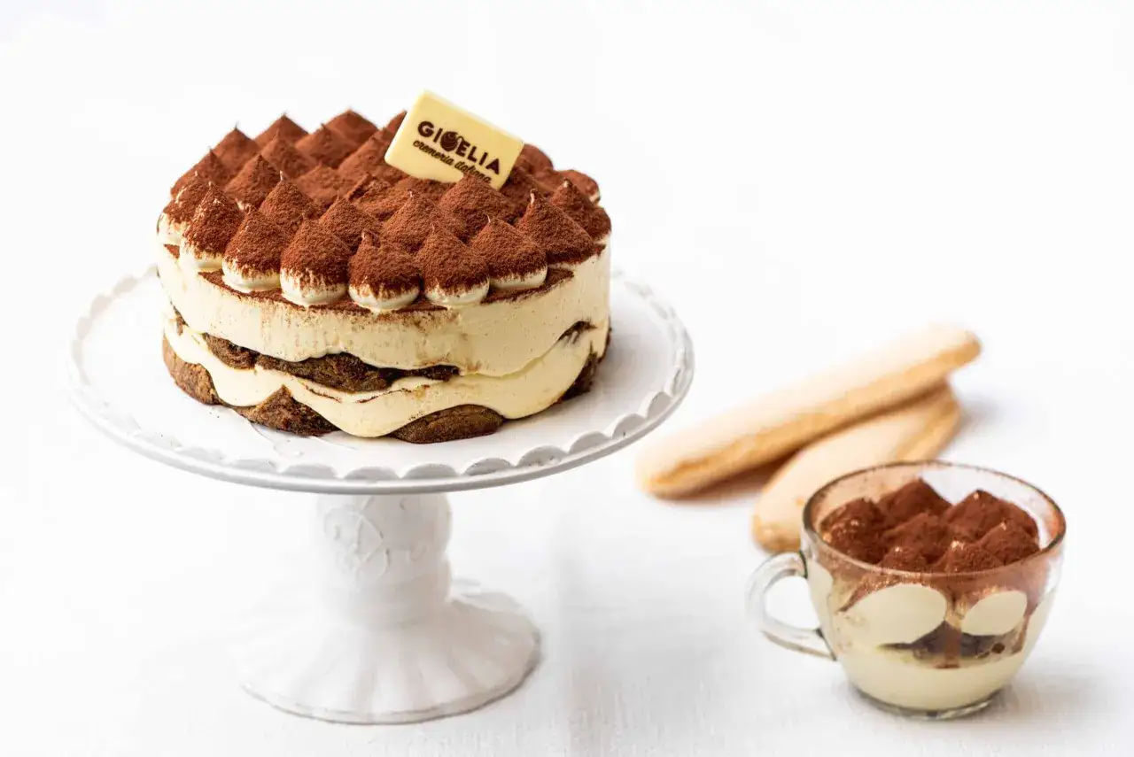 Un délicieux tiramisu, rappelant son origine italienne, est présenté sur un plat blanc, accompagné de biscuits à la cuillère et d'une tasse individuelle.
