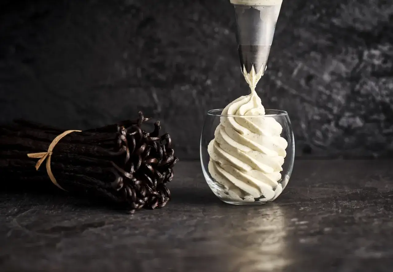 Une poche à douille dépose une crème diplomate vanillée dans un verre. Des gousses de vanille sont à côté, suggérant la recette.