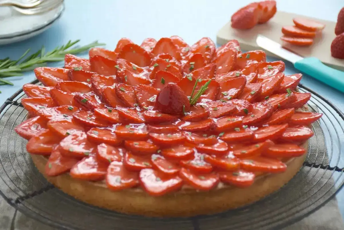 Tarte aux fraises magnifiquement décorée, avec un nappage fraise brillant et des éclats de romarin. Un délice visuel et gustatif.