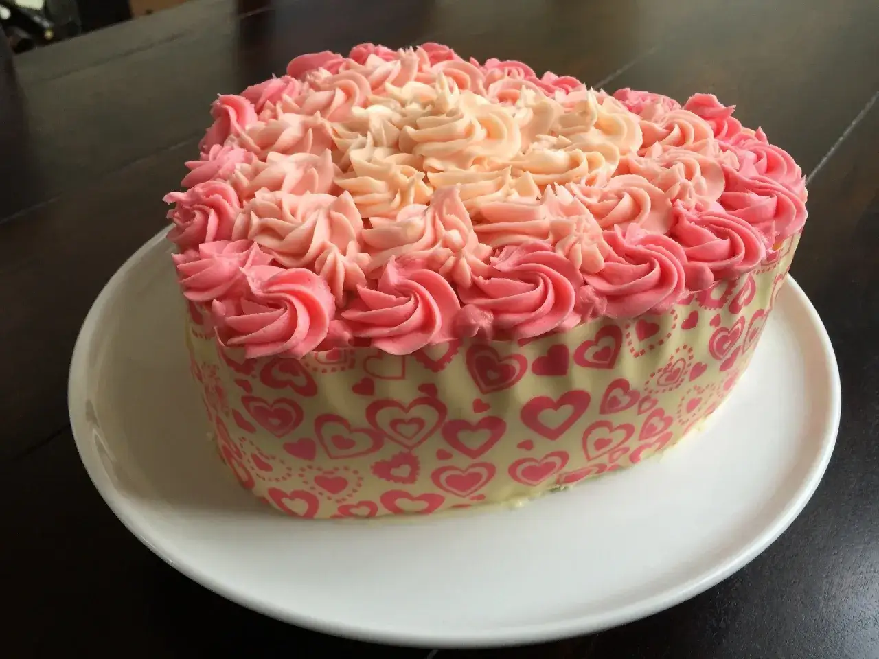 Gâteau en forme de cœur décoré de crème au beurre facile rose et pêche, entouré d'une bande de chocolat blanc ornée de cœurs roses.