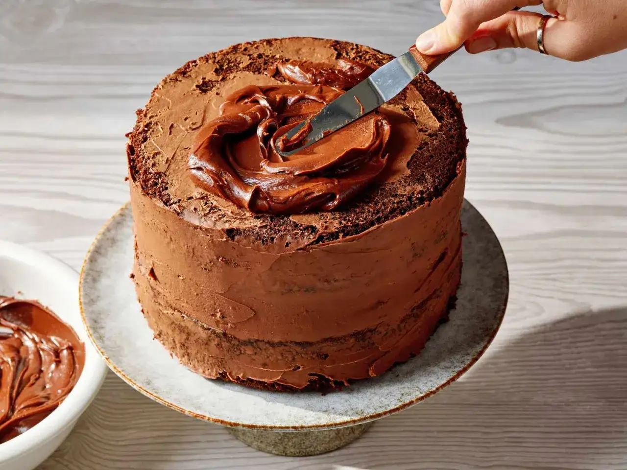 Une main étalant une délicieuse crème de couverture sur un layer cake au chocolat. Le glaçage est onctueux et appétissant.