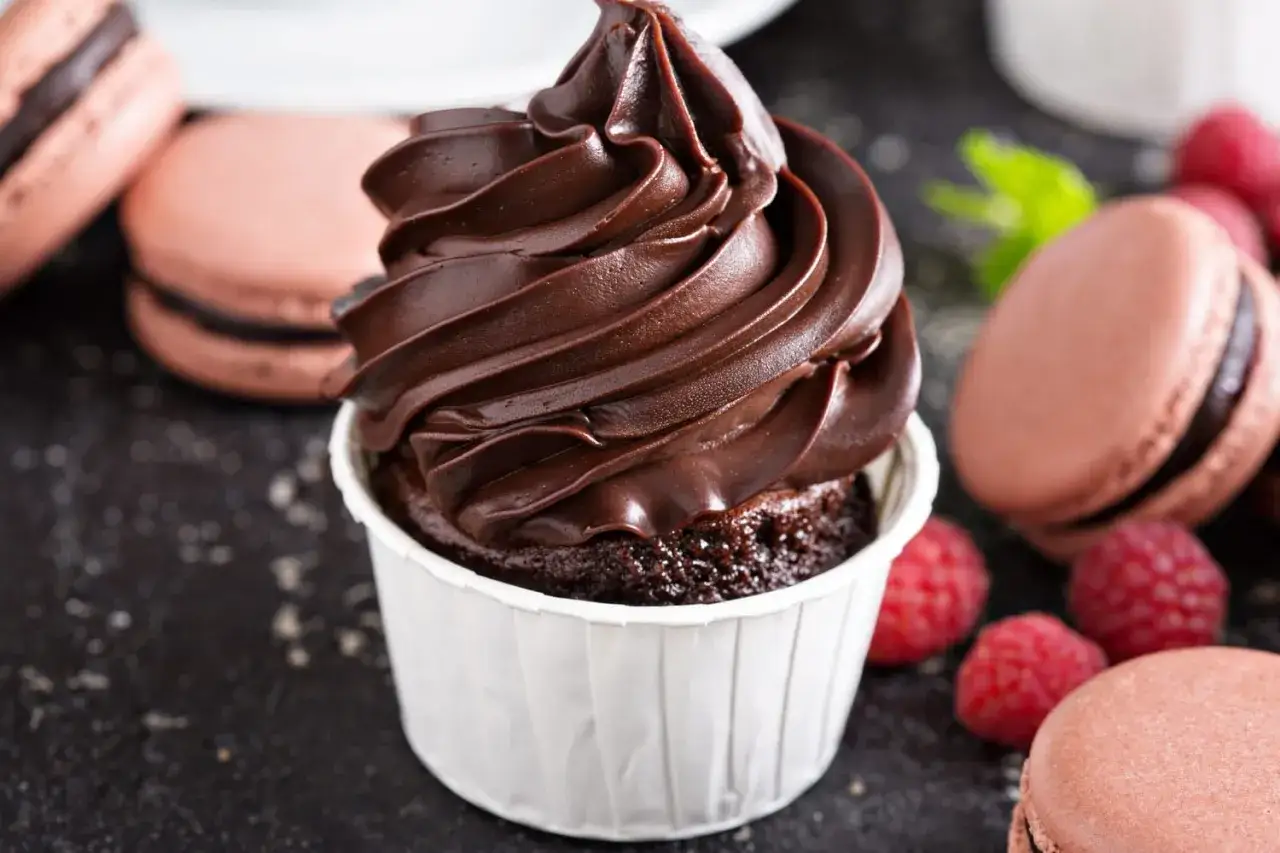 Un cupcake au chocolat avec une généreuse ganache chocolat, entouré de macarons roses et de framboises fraîches.