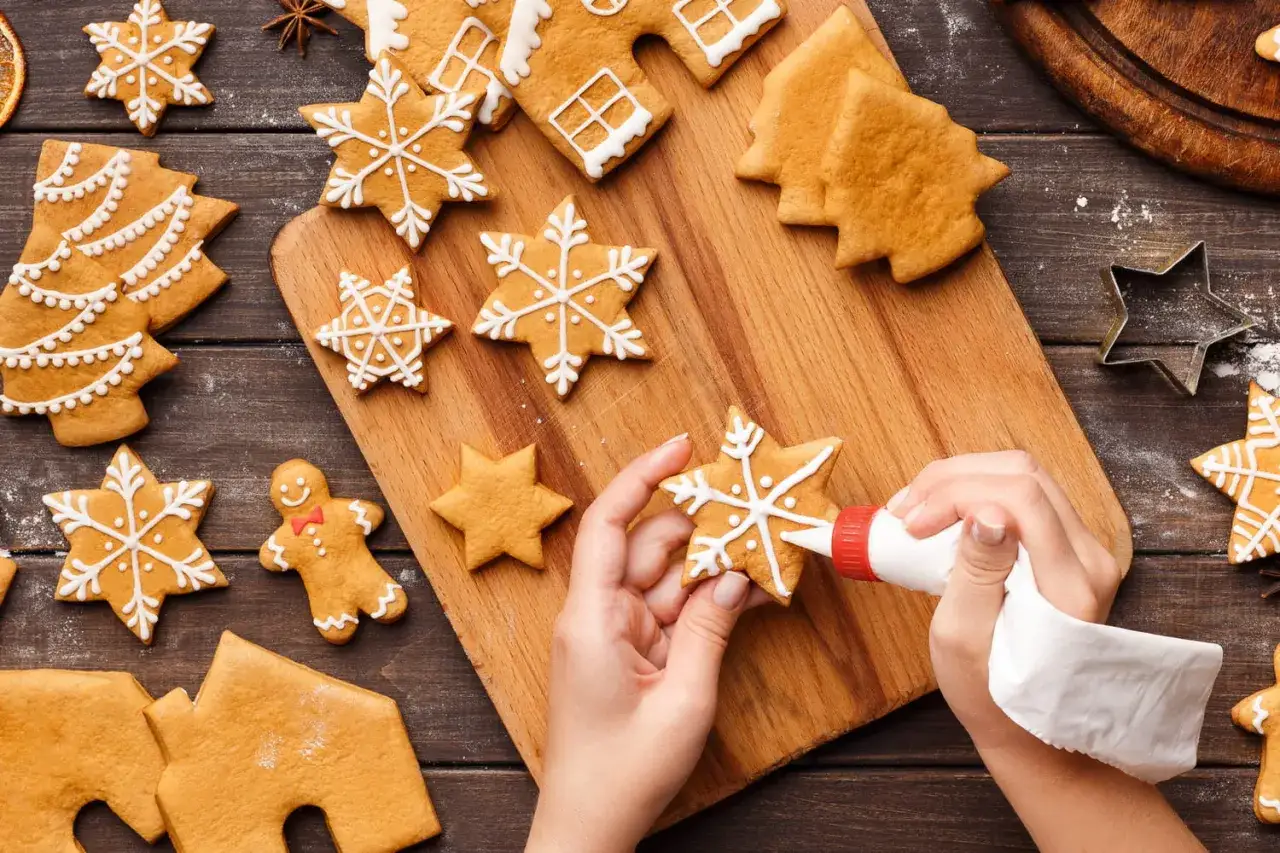 Des mains décorent un biscuit de noël en forme d'étoile avec du glaçage blanc. D'autres biscuits de noël, dont des sapins et des bonshommes, sont disposés autour.