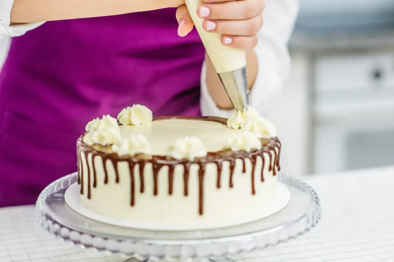 Une main décore un gâteau avec un glaçage crémeux, ajoutant des touches finales à un simple cake design avec un glaçage au chocolat dégoulinant.