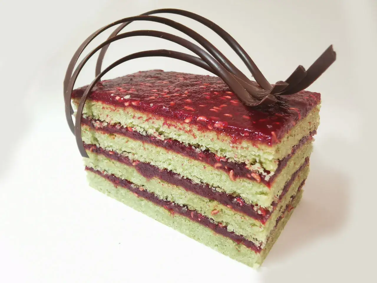 Tranche de layer cake pistache framboise, garnie de framboises et décorée de chocolat.