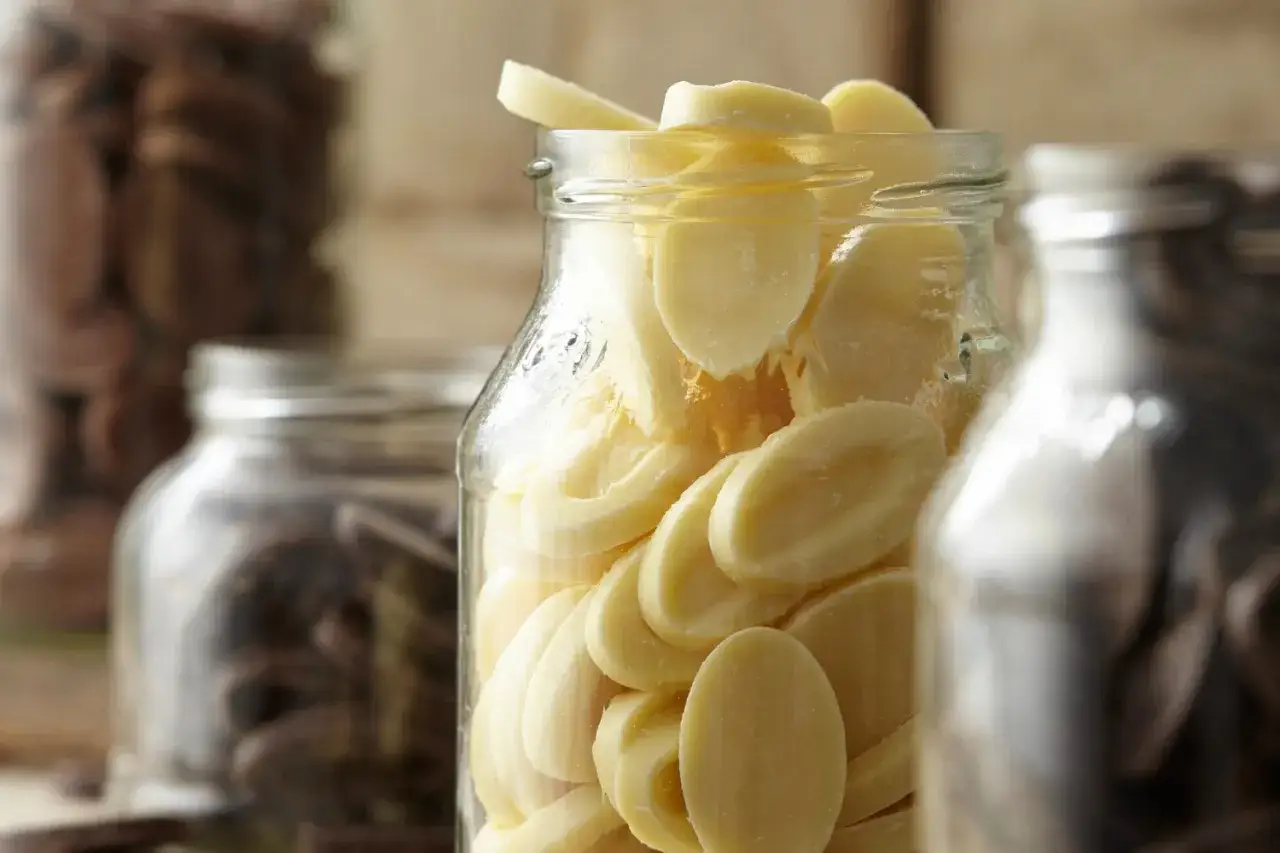 Des éclats de chocolat blanc dans un bocal, prêts pour la conservation du chocolat maison. Des fèves de cacao dans d'autres bocaux en arrière-plan.