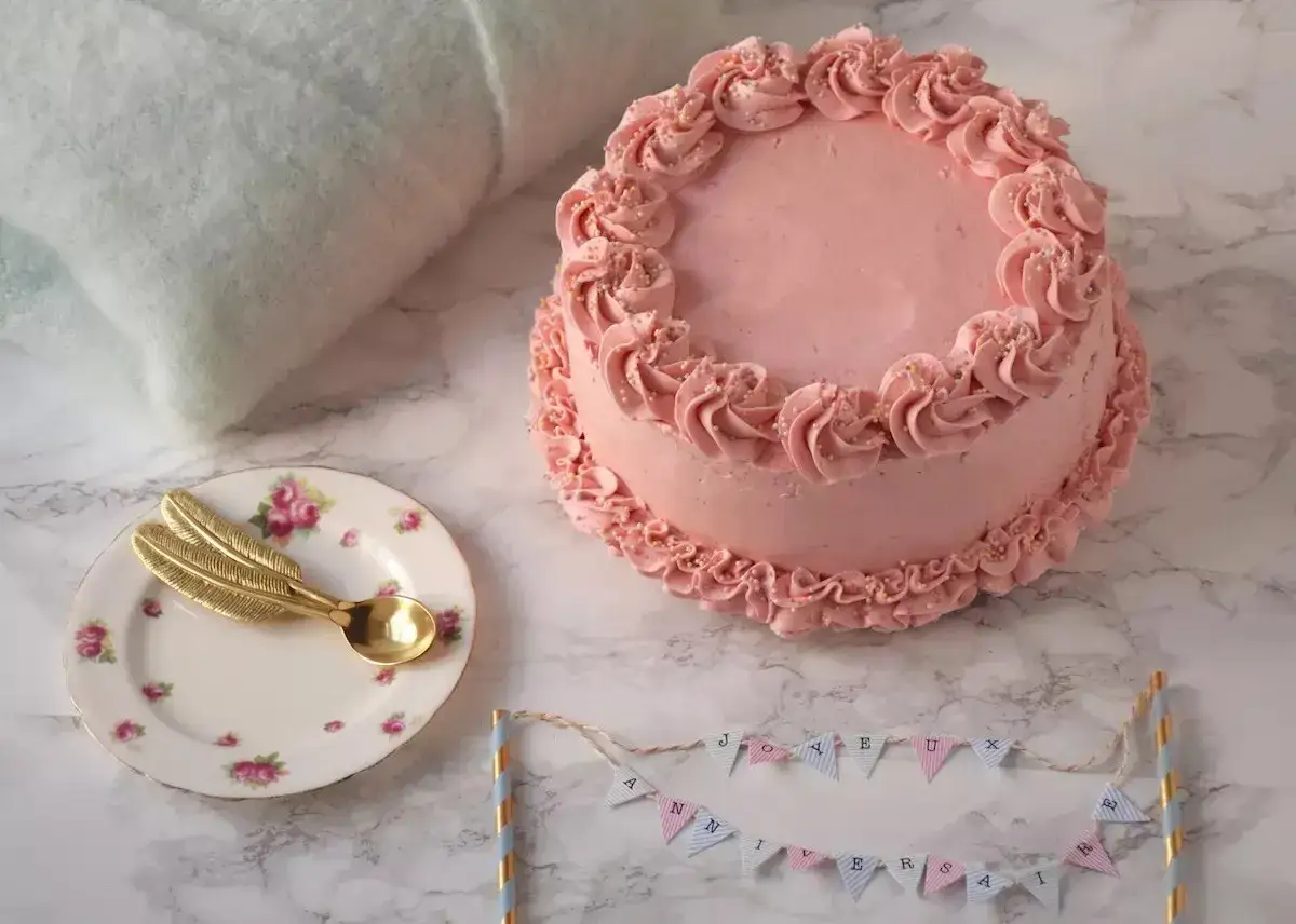 Gâteau rose avec un simple cake design, décoré de volutes et de perles. Une petite assiette avec une cuillère dorée et une guirlande