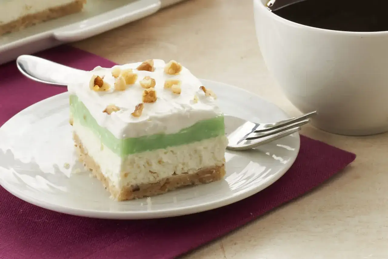 Délicieux entremet pistache à trois couches, garni de crème fouettée et de noix, servi sur une assiette blanche avec une tasse de café.