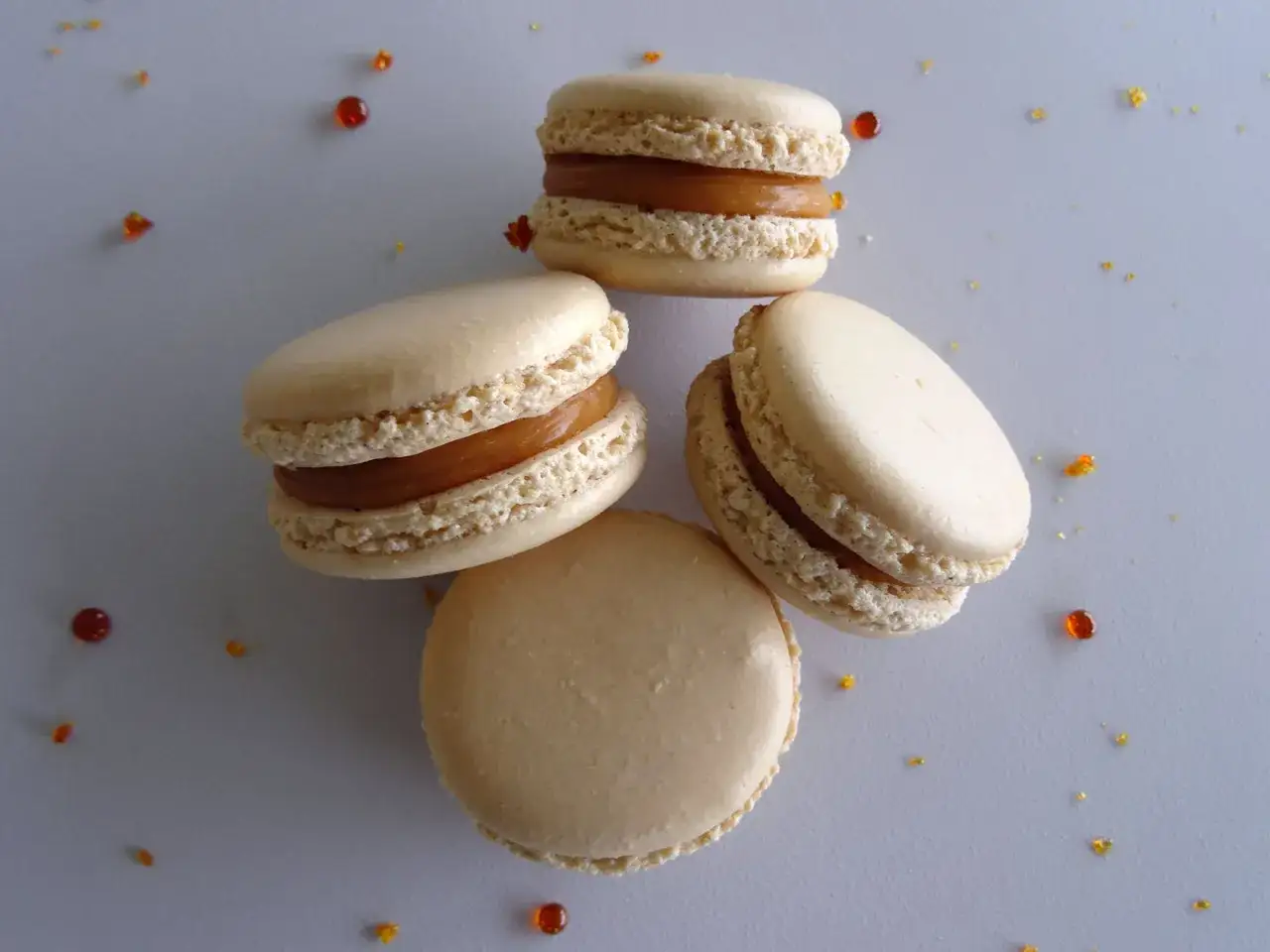 Quatre macarons clairs garnissent un fond gris, parsemé de quelques gouttes de caramel au beurre salé.