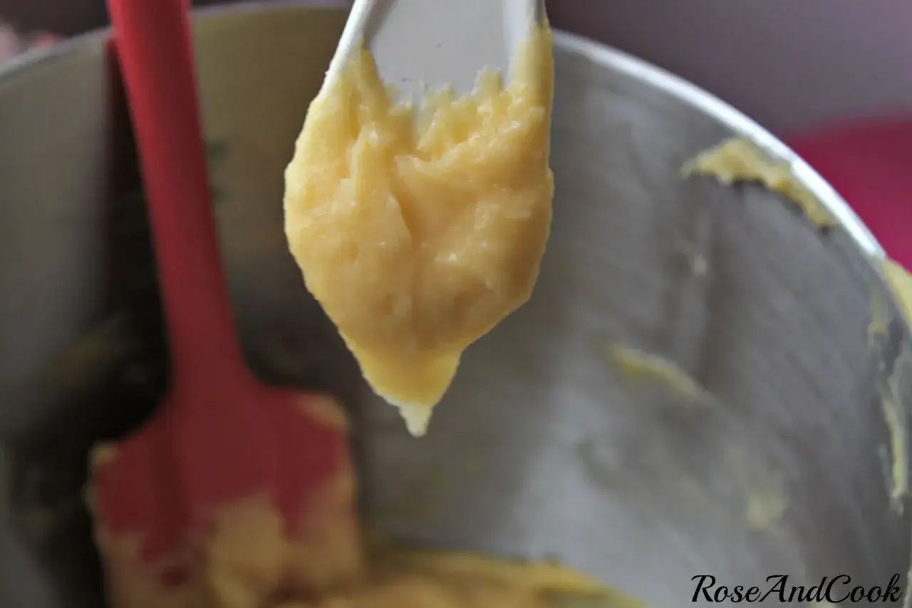 Une cuillère tient une pâte à choux trop liquide qui coule. Une spatule rouge est visible à gauche.