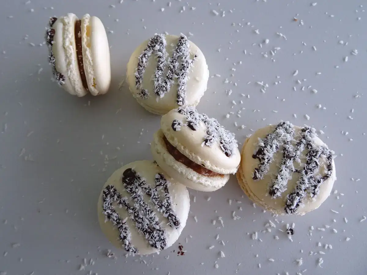 Macarons blancs garnis d'une ganache coco et saupoudrés de noix de coco râpée et de copeaux de chocolat.